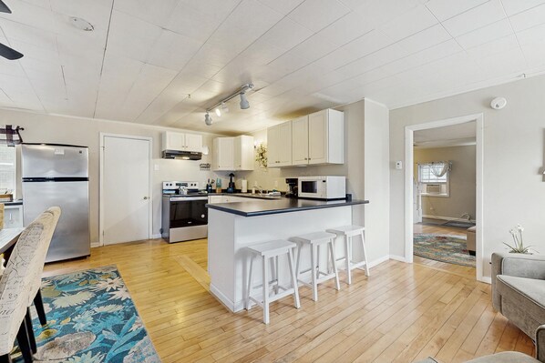 A modern kitchen with white cabinets, a stainless steel refrigerator, a built-in oven, and a microwave. There is a wooden dining table with chairs and a patterned rug.