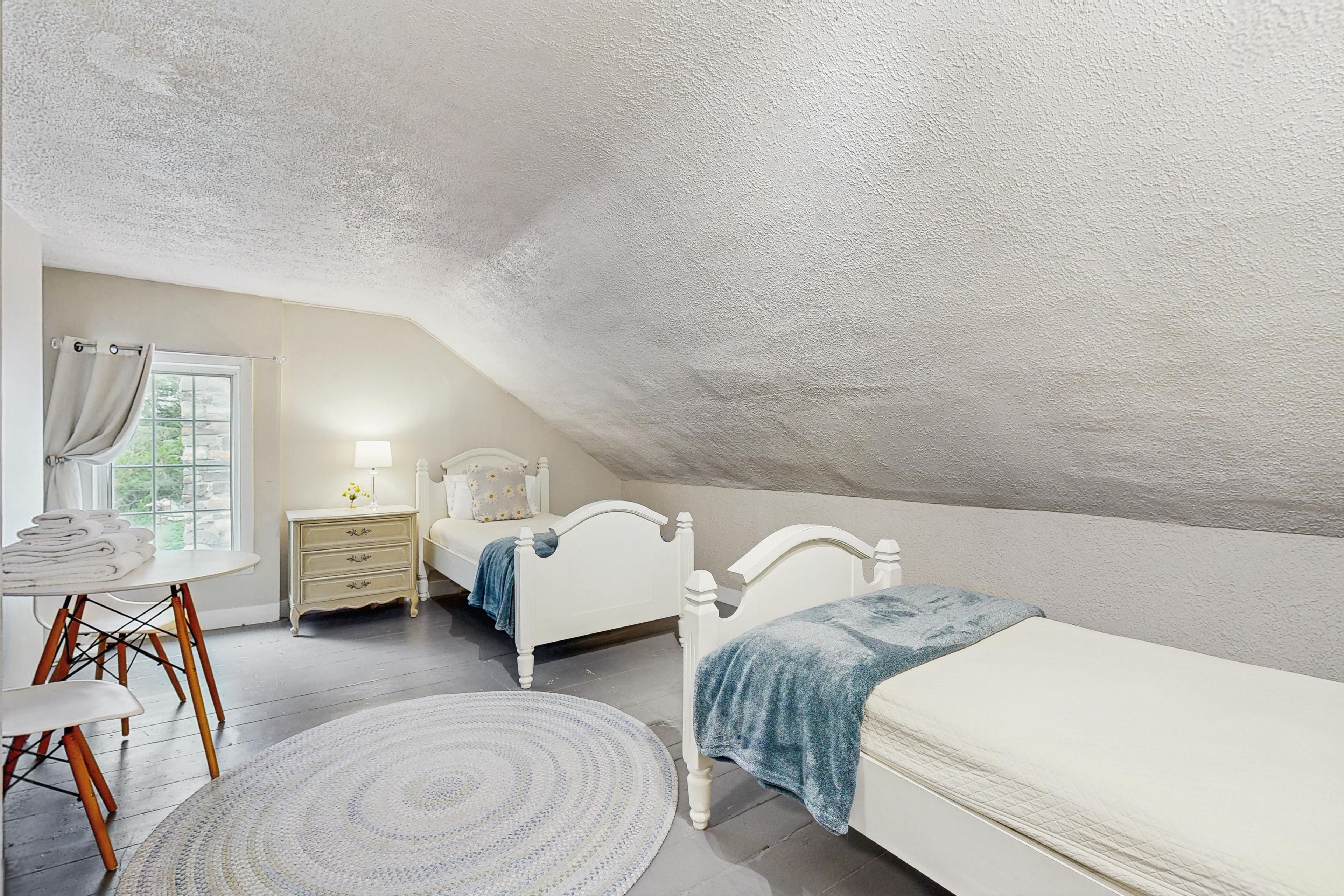 Un dormitorio con techo inclinado, una cama, una mesita de noche con lámpara, una silla y un ventanal con cortinas.