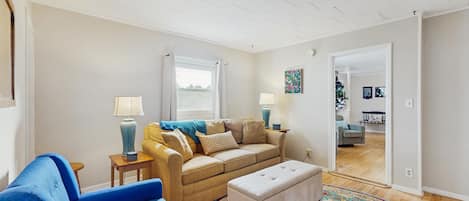 Ein Wohnzimmer mit einer Couch, einem blauen Sessel, einem gemusterten Teppich und einem Fenster mit Vorhängen.