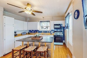 Une cuisine avec des armoires blanches, un îlot central en bois doté de tabourets, un ventilateur de plafond et un pare-feu en carreaux bleus.