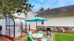 Ein überdachter Patio-Bereich mit Tisch und Stühlen, einem Grill und einem Gewächshaus.