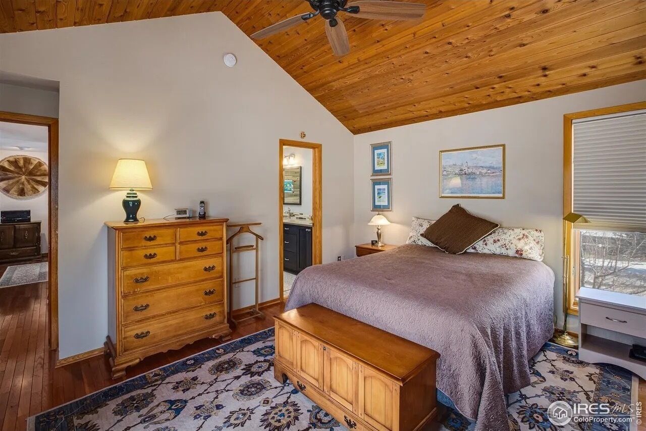 A bedroom with a wooden ceiling, a bed with a headboard, a dresser, a nightstand, a lamp, a mirror, and framed pictures on the wall.