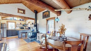 A rustic cabin interior with a wooden dining table, chairs, a kitchen area, and a fireplace. - Renovated historic cabin with mountain views, lake access, & electric fireplace (Jay)