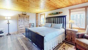 A bedroom with a wooden ceiling, a bed with white bedding, a nightstand, a chair, and a dresser. - Renovated historic cabin with mountain views, lake access, & electric fireplace (Jay)