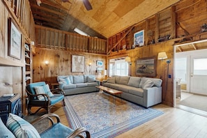 A cozy living room with wooden walls, a large sofa, a patterned rug, and a dining table. - Lakeside cottage w/ mountain views, canoes, firepit, enclosed porch, bbq, beach (Colchester)
