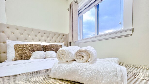 A neatly made bed with rolled white towels, a tufted headboard, and a window with a view of the sky. - *AC/8mins-YVR/Private Bathroom Suite/Separate Entry/Free Parking/Near Skytrain (Richmond)