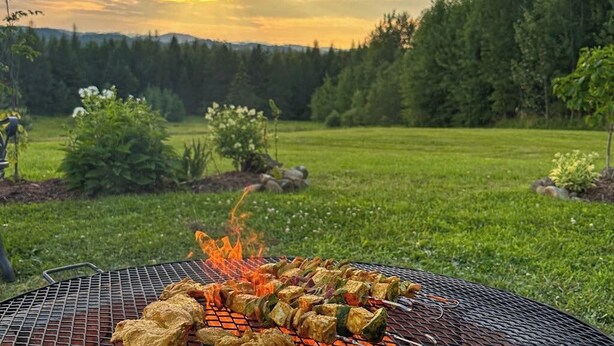 A barbecue grill with food and flames, set in a grassy outdoor area with trees in the background. - Cozy house w/ mountain views, large yard, firepit, wifi, jetted tub, TV, EV (Glover)
