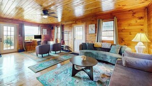 A wooden cabin interior with a sofa, chairs, a coffee table, and a television. - Cozy house w/ mountain views, large yard, firepit, wifi, jetted tub, TV, EV (Glover)
