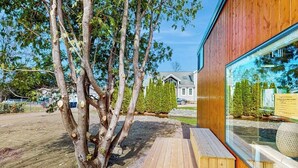 Un edificio moderno con una terraza de madera, un ventanal grande y un árbol en primer plano.