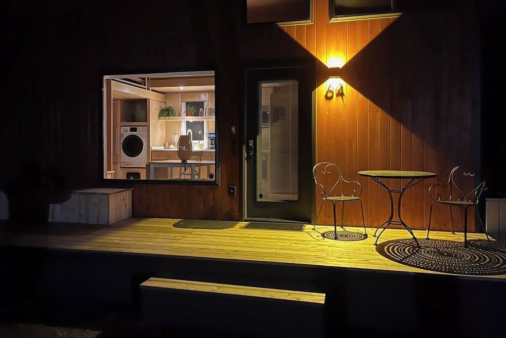 A wooden exterior with a door, a small outdoor seating area with two chairs and a table, and a visible interior with a washing machine and kitchen area.