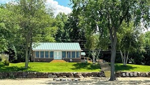 A house with a green roof, a stone wall, and a dock leading to a body of water. - Lakefront house w/ views, beach, fireplace, firepit, central air, arcade games (Colchester)