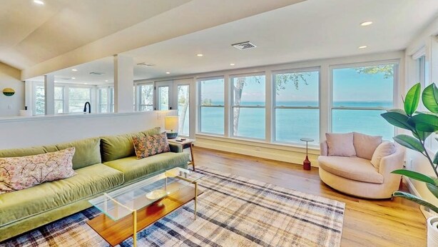 A spacious living room with a large window overlooking a body of water, a green sofa, a beige armchair, and a coffee table. - Lakefront house w/ views, beach, fireplace, firepit, central air, arcade games (Colchester)