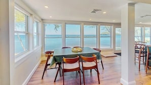 A dining area with a table and chairs, large windows, and a view of the sea. - Lakefront house w/ views, beach, fireplace, firepit, central air, arcade games (Colchester)