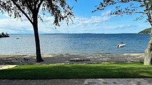 A lakeside view with a boat, grassy area, and a paved walkway. - Lakefront house w/ views, beach, fireplace, firepit, central air, arcade games (Colchester)