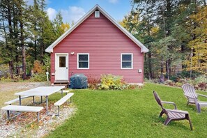 A red wooden cabin with a white door, two windows, a picnic table, and two Adirondack chairs in a grassy yard surrounded by trees. - Cozy cabin close to Smuggler's Notch w/ firepit, gas grill, trail to river, wifi (Jeffersonville)