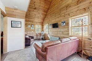 Een interieur van een blokhut met houten wanden, een roze leren bank, een eettafel met stoelen en een aan de muur bevestigde televisie.
