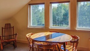 A room with a round wooden table, four chairs, and large windows with a view of trees.