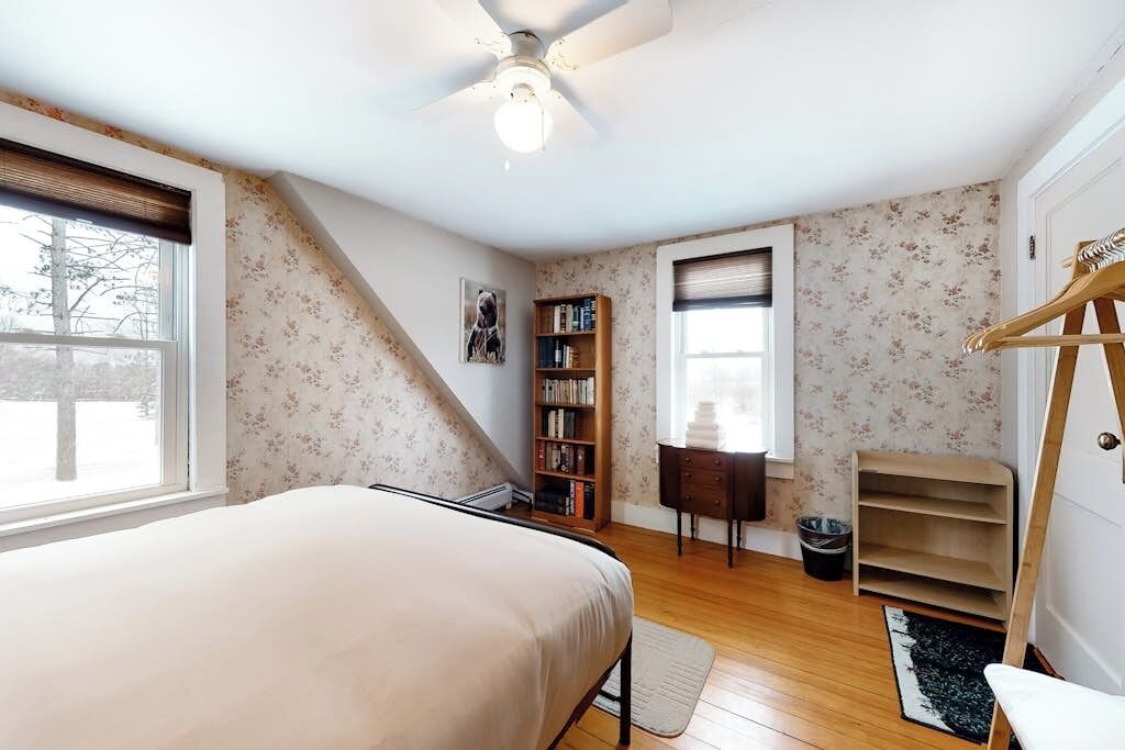 Un dormitorio con cama, ventilador de techo, estantería, escritorio, silla, perchero y ventana.