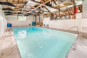 Une piscine intérieure dotée d’un sol en béton, de poutres en bois et d’un mur blanc percé d’une fenêtre.