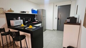 Una cucina compatta con forno a microonde, macchina del caffè e frigorifero. Sono presenti due sgabelli neri e un'isola con una ciotola gialla.