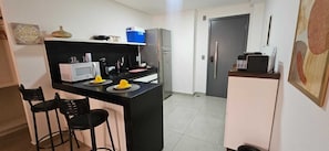 A compact kitchen with a microwave, coffee maker, and a refrigerator. There are two black bar stools and a small island with a yellow bowl. - Fully furnished apartment in João Pessoa, very close to the beach, with leisure area and garage. (João Pessoa)