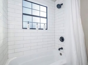 Un baño moderno con paredes de azulejos blancos, una ventana, una bañera y un cabezal de ducha.