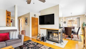 Una sala de estar con chimenea, televisor, comedor con candelabro y sofá con un cojín rojo.