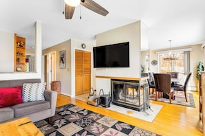 A living room with a fireplace, a television, a dining area with a chandelier, and a sofa with a red pillow. - Beautiful Stowe getaway w/ shared pool/hot tub, balcony & wood-burning fireplace (Stowe)
