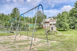 A playground with swings and a slide, surrounded by trees and a building. - Gorgeous Home with Free WiFi, Electric Fireplace, & Shared Pool/Hot Tub (Stowe)