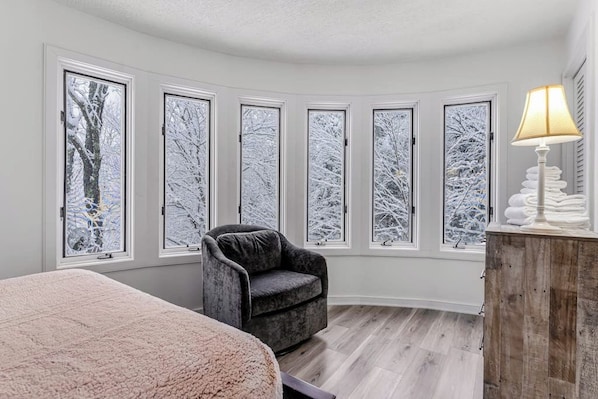 A bedroom with a large bed, a chair, a wooden cabinet, and a window with a view of snow-covered trees. - Three-Level House Near Skiing w/Free WiFi, Private Hot Tub, Gas Fireplace (Stowe)