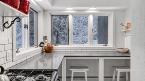 Ett kök med en gasspis, vita skåp, ett fönster med utsikt över snötäckta träd och en liten köksö med barstolar.