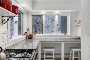 A kitchen with a gas stove, white cabinets, a window with a view of snow-covered trees, and a small island with stools. - Three-Level House Near Skiing w/Free WiFi, Private Hot Tub, Gas Fireplace (Stowe)