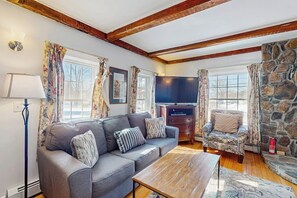 A living room with a sofa, armchair, wooden coffee table, stone wall, and a flat-screen TV. - Quaint Getaway Near Town W/ Hot Tub, Large Deck, Sauna & Free WiFi (Stowe)