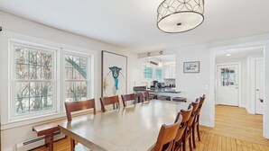 Un'area pranzo con un tavolo lungo e sedie, ampie finestre, una cucina sullo sfondo e una porta che dà su un'altra stanza.