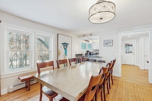 A dining area with a long table and chairs, large windows, a kitchen in the background, and a door leading to another room. - Quaint Getaway Near Town W/ Hot Tub, Large Deck, Sauna & Free WiFi (Stowe)
