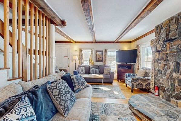 A cozy living room with a sofa, patterned cushions, a television, and a stone wall. - Quaint Getaway Near Town W/ Hot Tub, Large Deck, Sauna & Free WiFi (Stowe)