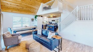 A spacious living area with a wooden ceiling, a staircase, a kitchen, and a seating area with a sofa and armchairs. - Waterfront home on Lake Champlain with pool, hot tub, dock, kayak & canoe (North Hero)