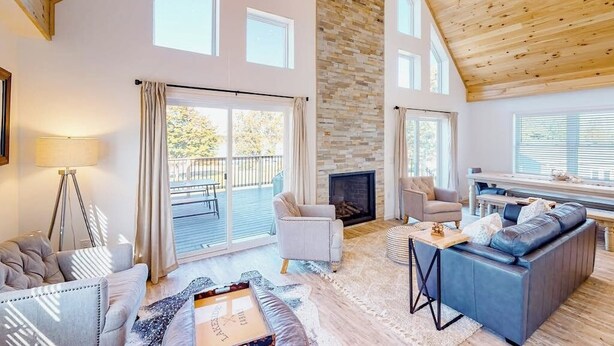 A spacious living room with a fireplace, large windows, and a high ceiling. - Waterfront home on Lake Champlain with pool, hot tub, dock, kayak & canoe (North Hero)