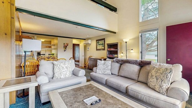 A modern living room with a sofa, coffee table, and a dining area with chairs. - Golf course-front condo with loft, indoor & outdoor pools, hot tub, & game room (Stowe)