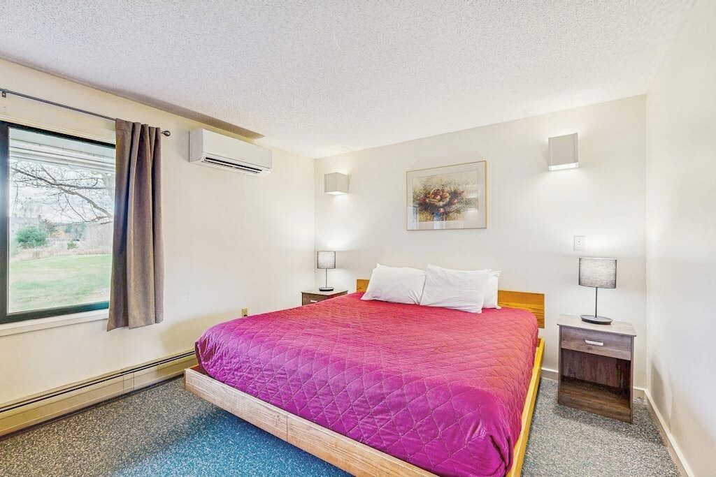 A hotel room with a bed, two bedside tables, a window with curtains, an air conditioning unit, and a wall-mounted artwork.