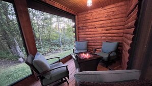A log cabin interior with a wooden deck, two chairs, a table with a fire, and a large window overlooking a forest. - Enchanting dog-friendly cabin with fireplace, hot tub & washer/dryer (Morrisville)