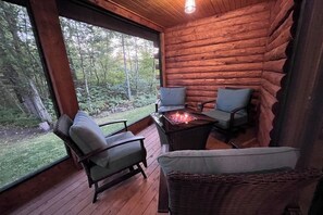 L’intérieur d’une cabane en rondins, avec une terrasse en bois, deux chaises, une table avec un foyer et une grande fenêtre donnant sur une forêt.
