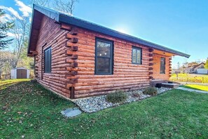 Une cabane en rondins avec un porche à l’avant, un petit jardin devant et un ciel dégagé.