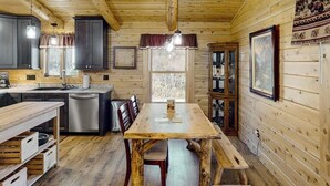 A wooden cabin interior with a dining table, chairs, a kitchen area, and a window. - Enchanting dog-friendly cabin with fireplace, hot tub & washer/dryer (Morrisville)