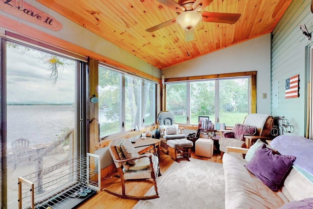 Une cabane avec un intérieur en bois, de grandes fenêtres, un ventilateur de plafond et une vue sur une étendue d’eau.
