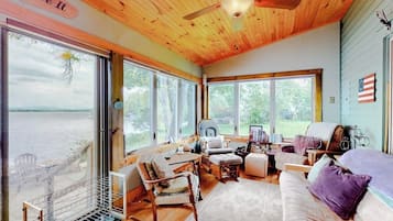 Une cabane avec un intérieur en bois, de grandes fenêtres, un ventilateur de plafond et une vue sur une étendue d’eau.