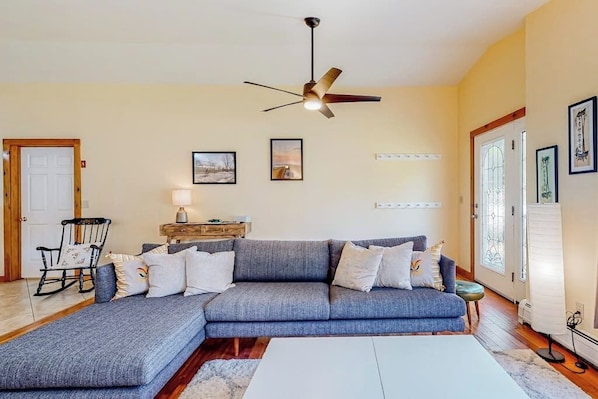 A living room with a sectional sofa, a rocking chair, a ceiling fan, and framed pictures on the wall.
