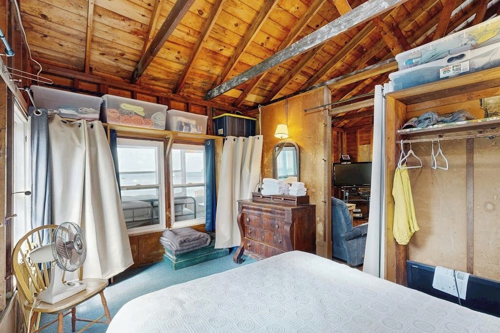 Interior de una cabaña de madera con una cama, una silla, un aparador y una ventana.