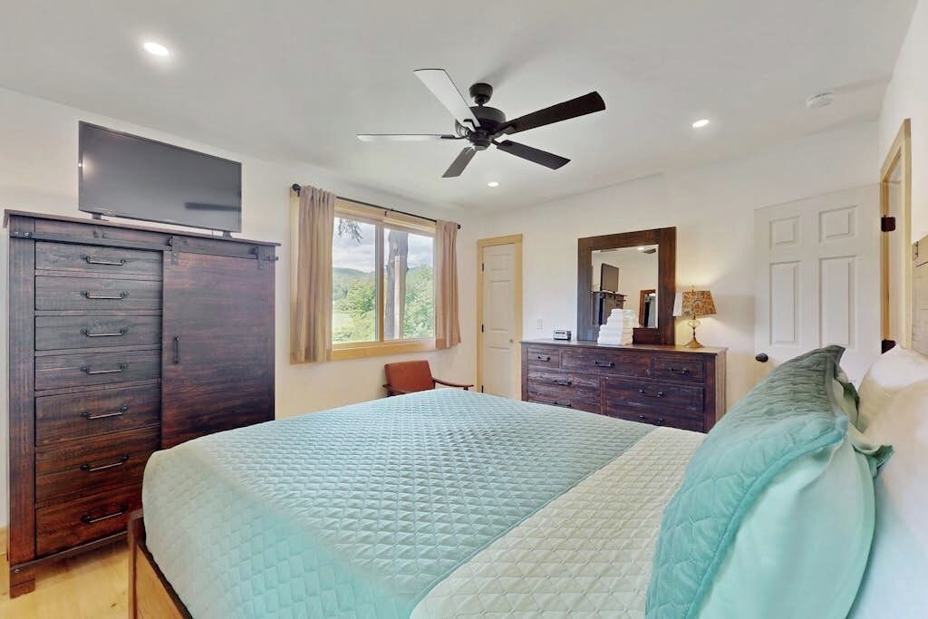 Un dormitorio con cama, ventilador de techo, televisor, cómoda y ventana con cortinas.