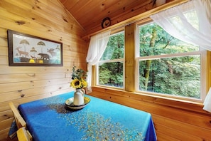 L’intérieur d’un chalet en bois, avec une grande fenêtre, un tableau encadré représentant des animaux sauvages, et une table recouverte d’une nappe bleue agrémentée d’un arrangement de tournesols.